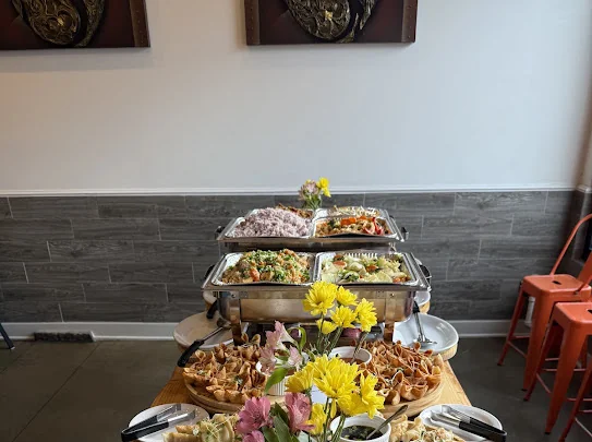 Catering buffet from Aloy Thai Eatery in Cherry Hills Village with chafing trays of rice and stir-fried dishes behind platters of appetizers and a vase of yellow flowers