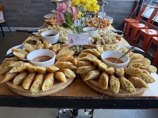 Two round wooden platters piled with golden fried egg rolls, each with a bowl of dipping sauce, on a catering table with fresh flowers from Aloy Thai Eatery in Cherry Hills Village